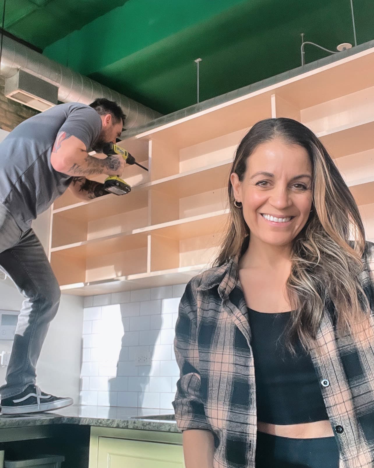 Jenn smiling while Shane installs shelving behind her, drill in hand