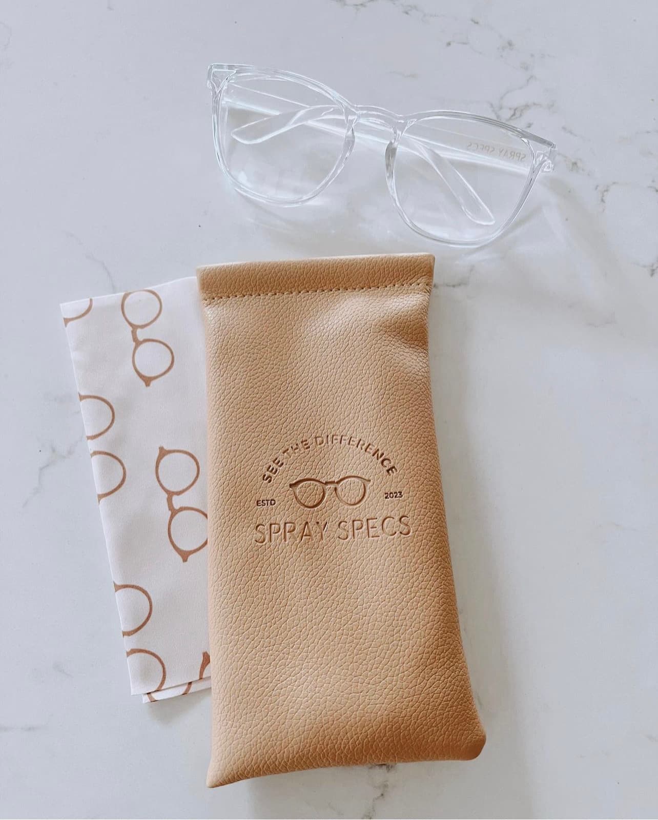 Spray Specs product flat-lay — clear glasses and embossed tan leather pouch on marble